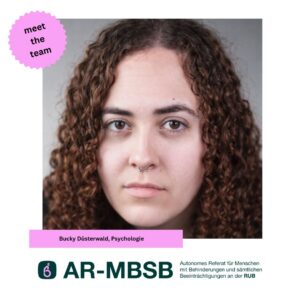 Bucky ist frontal vor neutralem Hintergrund porträtiert. Sie hat schulterlanges, lockiges braunes Haar, braune Augen und trägt ein Septum-Piercing. Sie blickt ruhig und ernst in die Kamera.